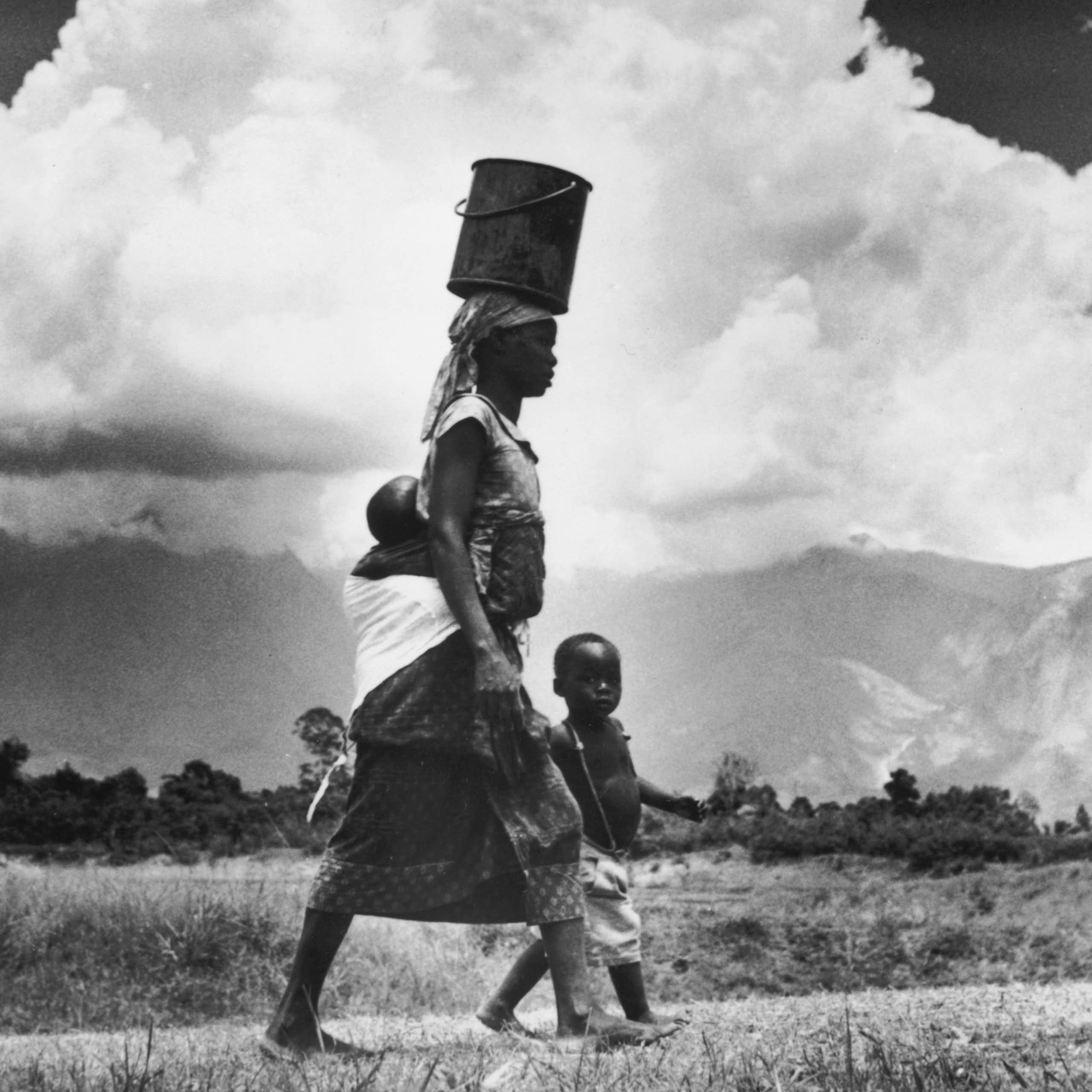 woman walks with water bucket on her head, baby strapped to her back and a young child at her side