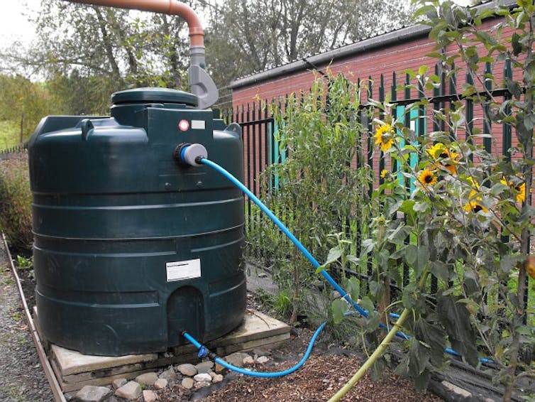 A large tank with an intake pipe at the top and pipes running from the bottom sits on a concrete slab in a garden next to a fence with wildflowers along it.