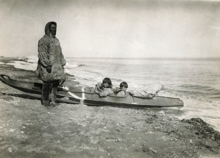 Man in traditional clothing stands on the bank next to a canoe with two children in it