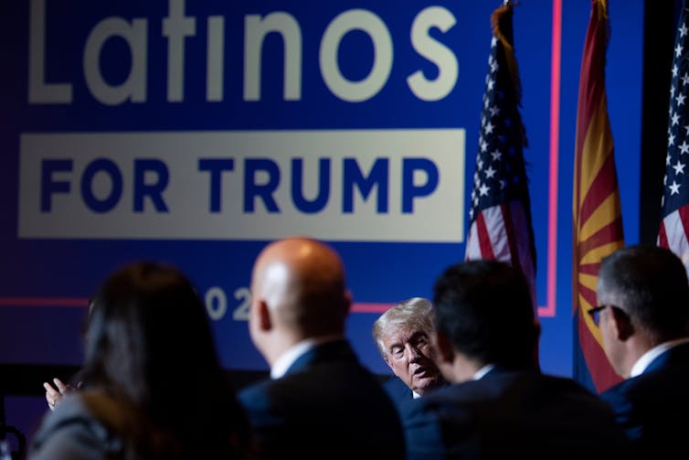Several men dressed in business suits stand in front of a large poster that reads Latinos for Trump.