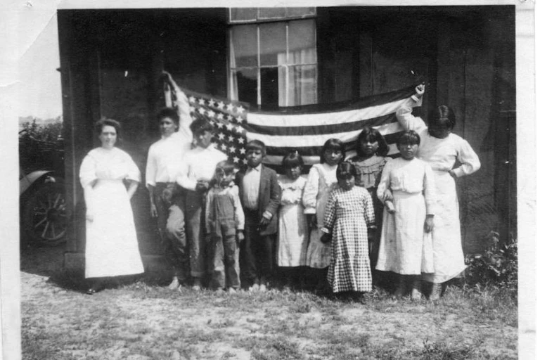 Black-and-white photo of young native children standing with an older white woman in front of an American flag and a dilapidated building.