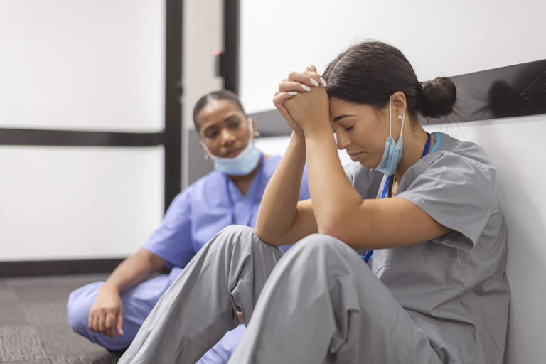 Two women with dark hair sit on the floor of a room with white walls, wearing blue and gray uniforms and looking exhausted.