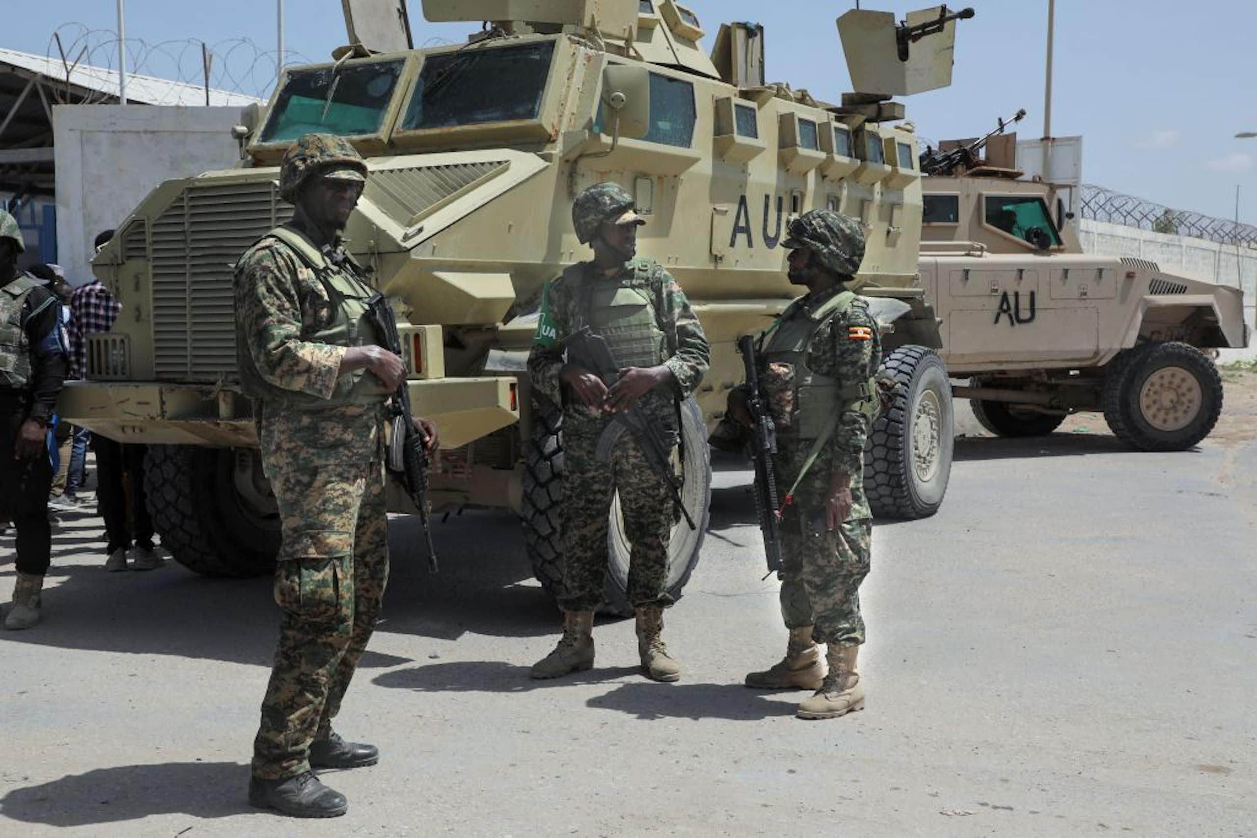 Three uniformed men holding guns standing in front of large, green armoured vehicles with the letters AU written in black on the sides. 