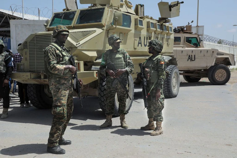 Three uniformed men holding guns standing in front of large, green armoured vehicles with the letters AU written in black on the sides.