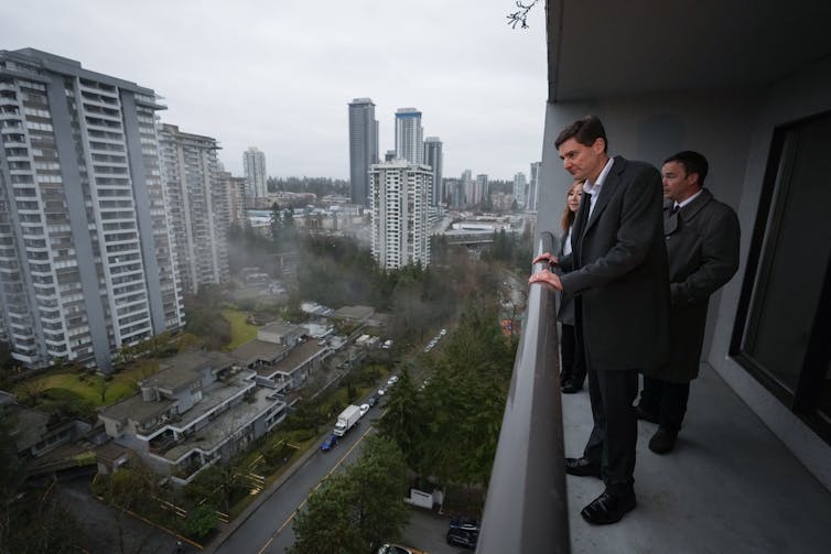 A man stands on the balcony of an apartment taking in a city view.