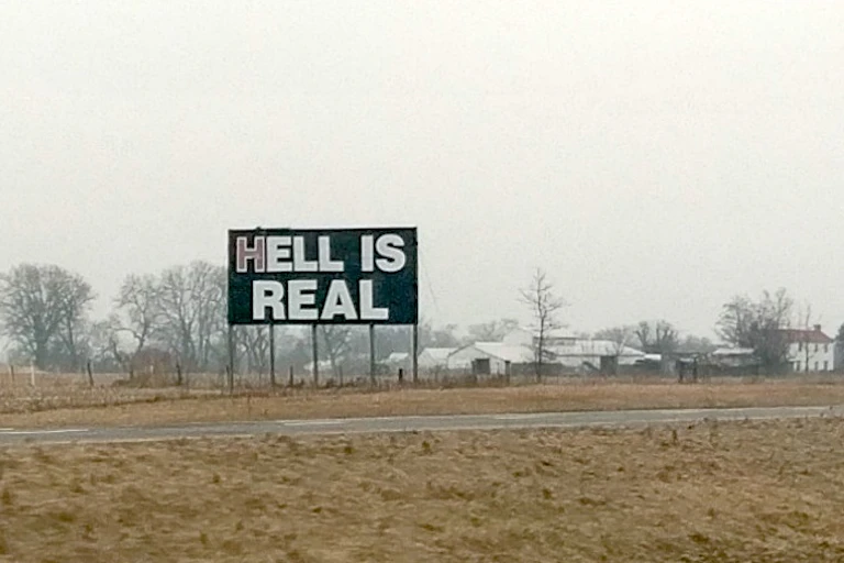 A sign saying 'Hell Is Real,' near a road with homes and trees in the background.