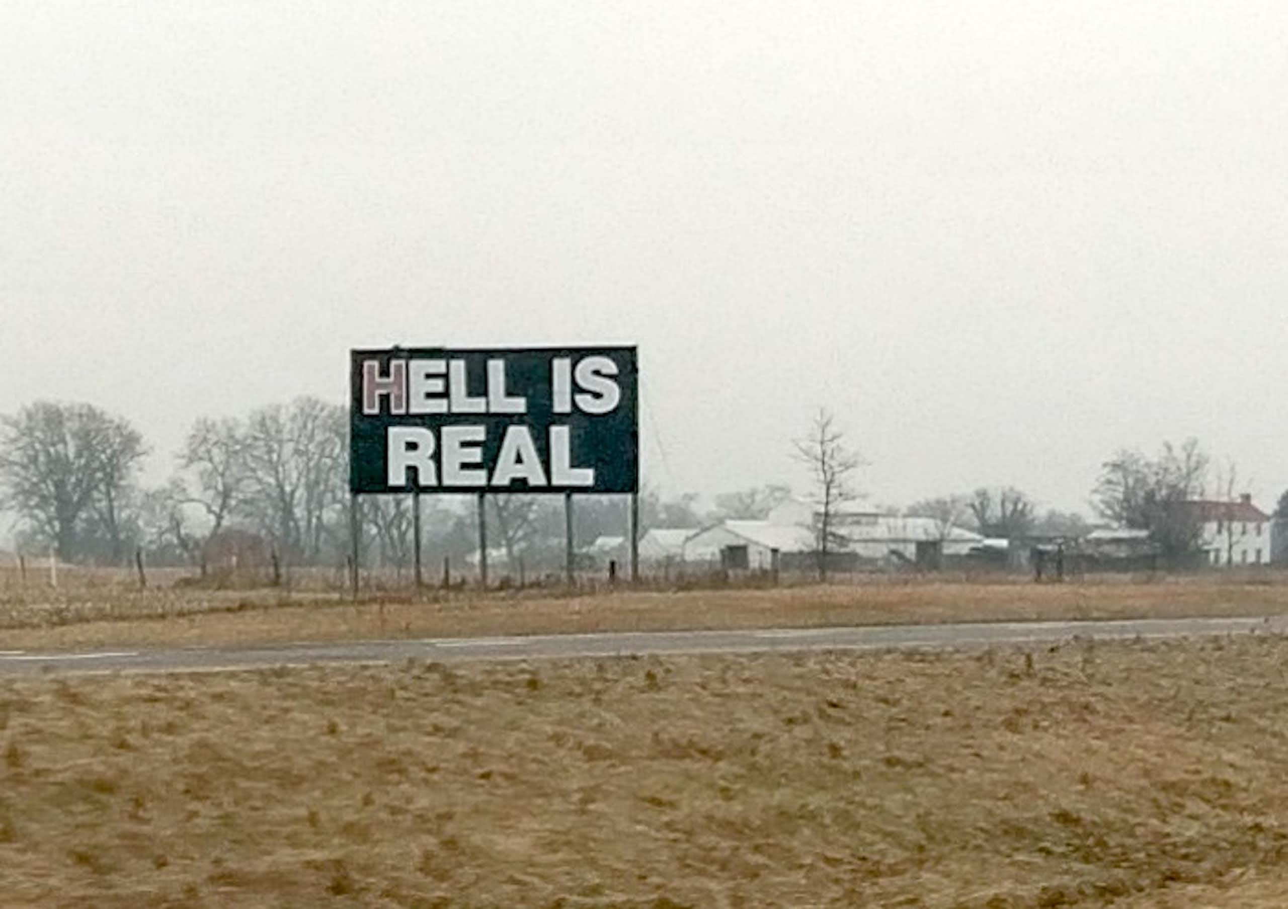 A sign saying 'Hell Is Real,' near a road with homes and trees in the background.