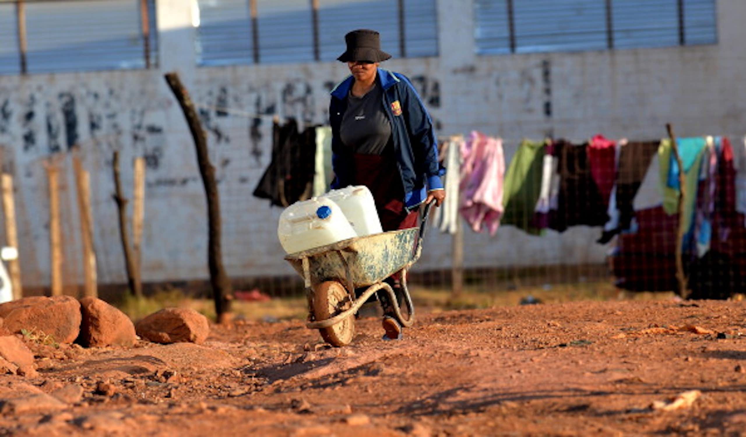 A woman pushes a wheelbarrow carrying containers of water. 