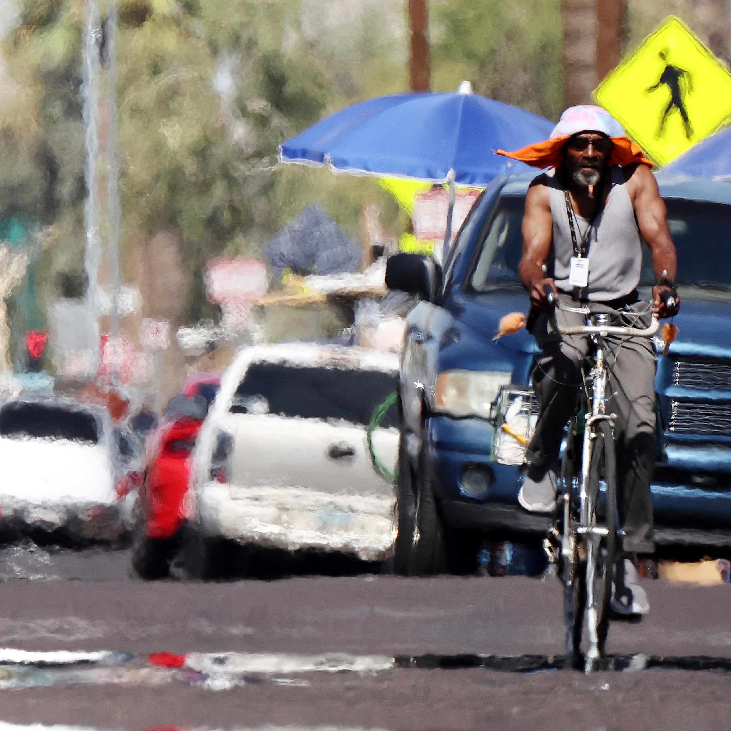 A man with a large hat rides a bike a on street with heat rising, creating a wavy view.