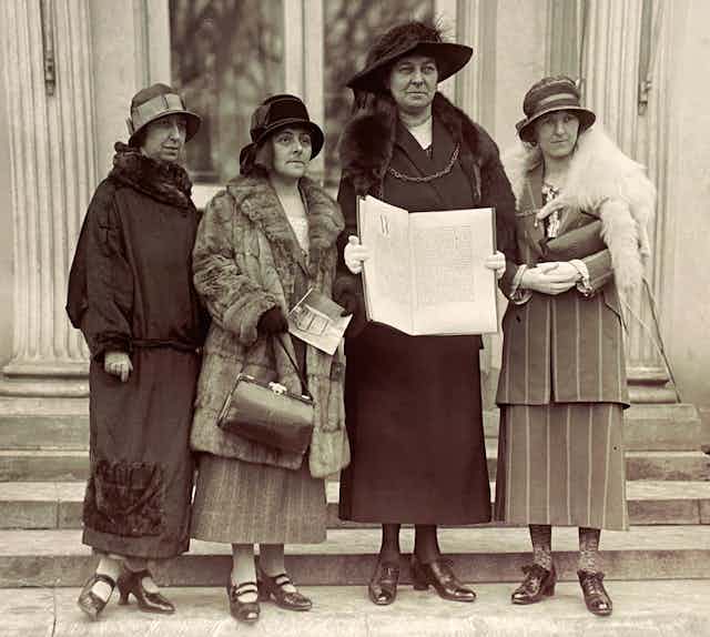 Four smartly dressed women with hats stand with a large book on the steps of the White House