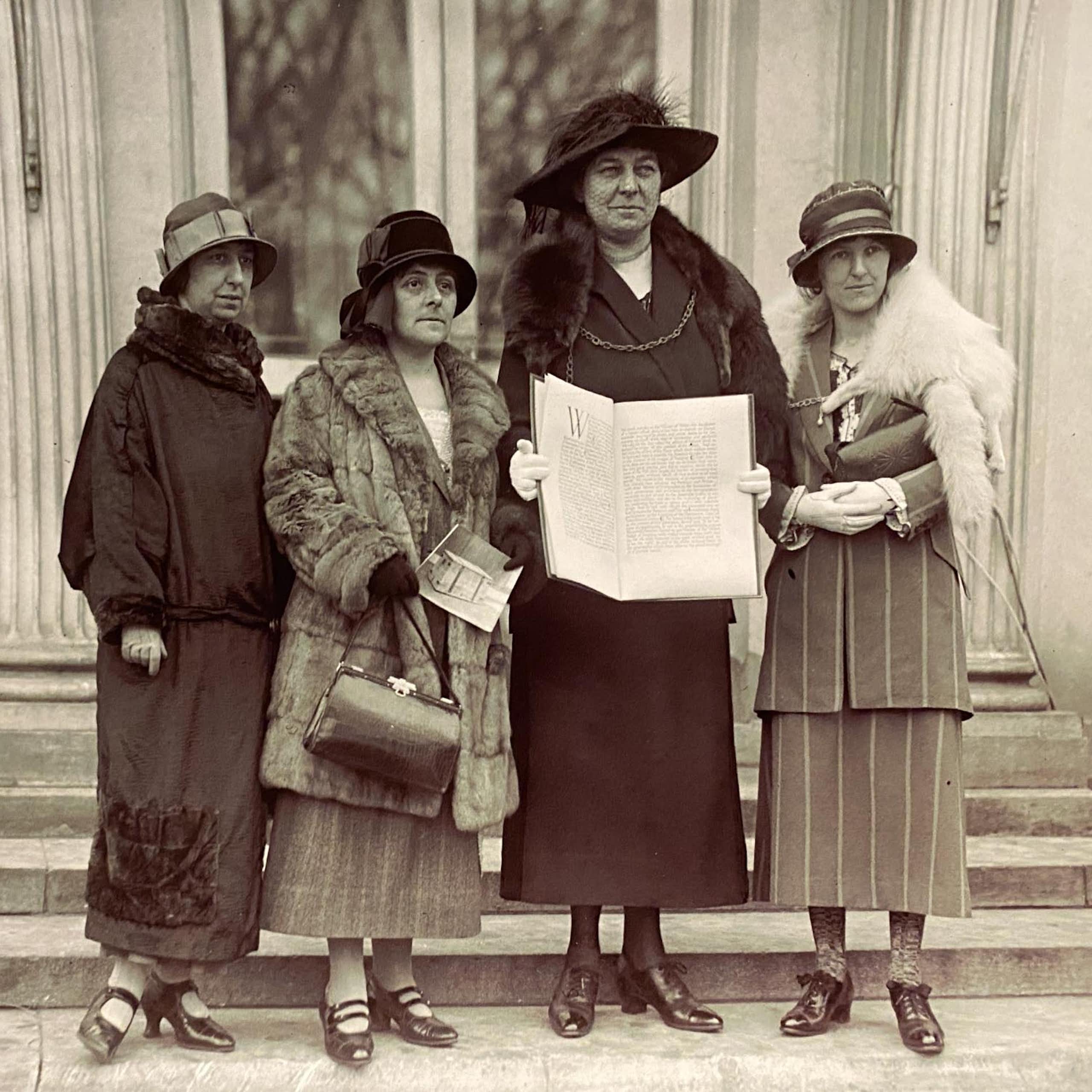 Four smartly dressed women with hats stand with a large book on the steps of the White House