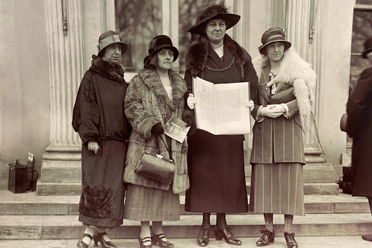Four smartly dressed women with hats stand with a large book on the steps of the White House