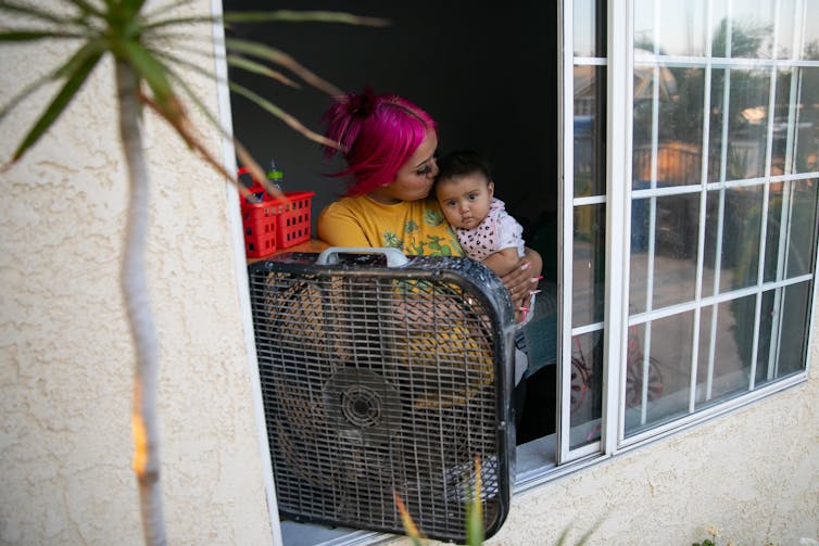 A woman holds a baby at an open window with a fan blowing in.