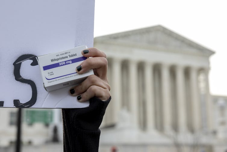 A hand with painted nails clutches a small package of medicine in front of a marble building with tall pillars.
