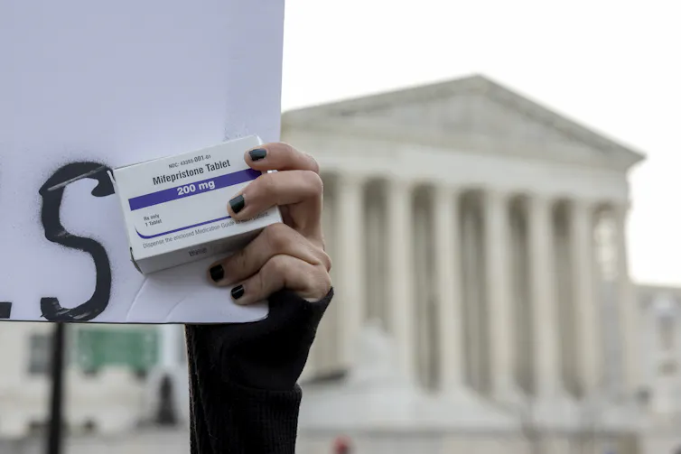 A hand with painted nails clutches a small package of medicine in front of a marble building with tall pillars.