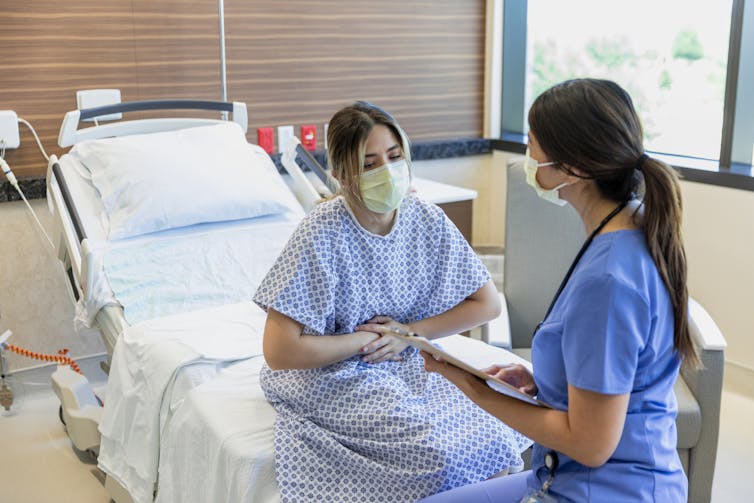 A woman in a medical mask and hospital gown sits on a bed and holds her stomach while talking to a woman in a blue gown.