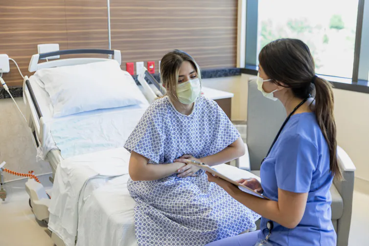 A woman in a medical mask and hospital gown sits on a bed holding her stomach as she speaks with a woman in blue scrubs.