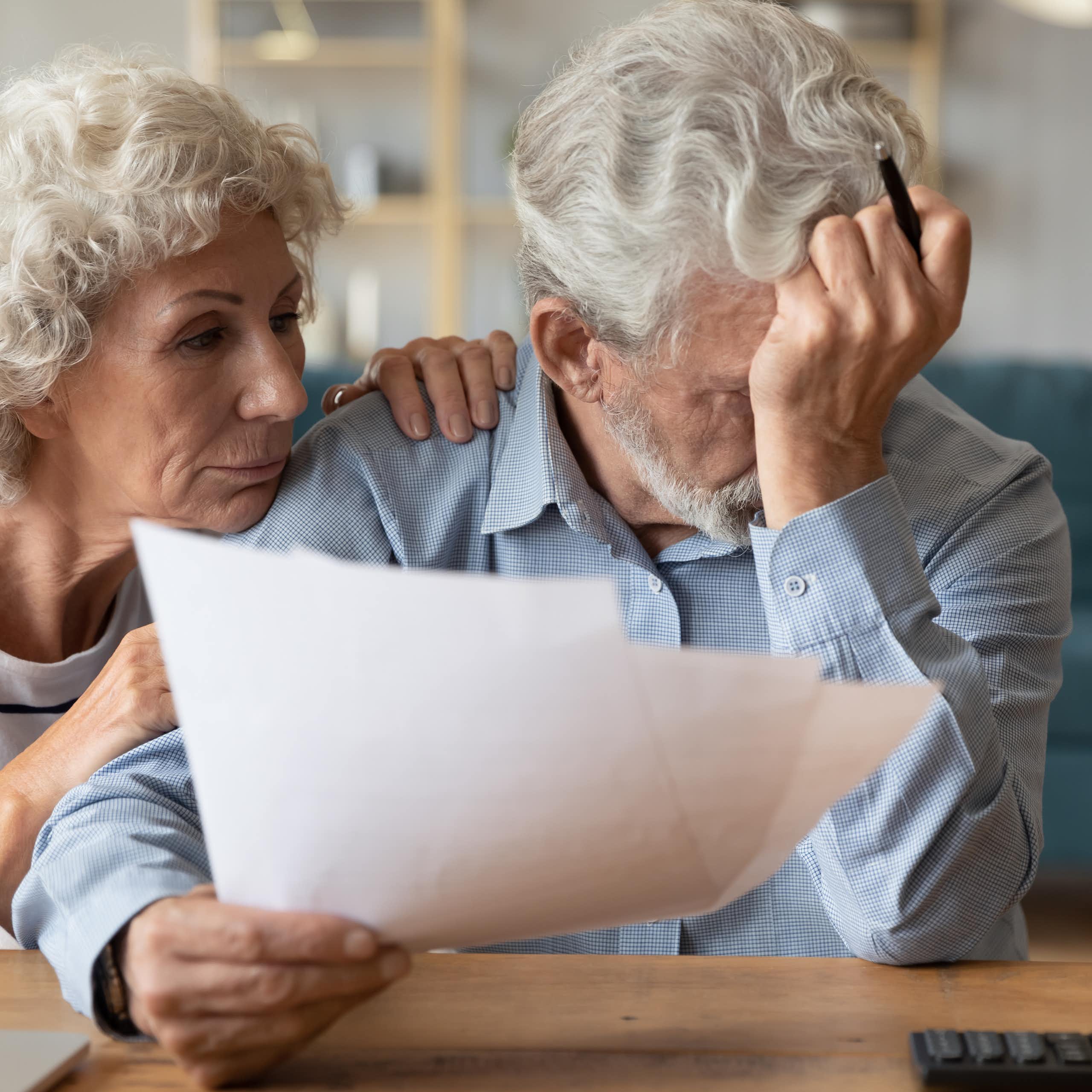 An older man holding a piece of paper and sadly resting his forehead on his hand while his wife, next to him, comforts him. There is a calculator on the table.