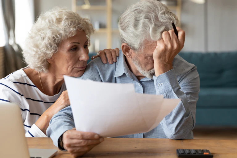 An older man holding a piece of paper and sadly resting his forehead on his hand while his wife, next to him, comforts him. There is a calculator on the table.