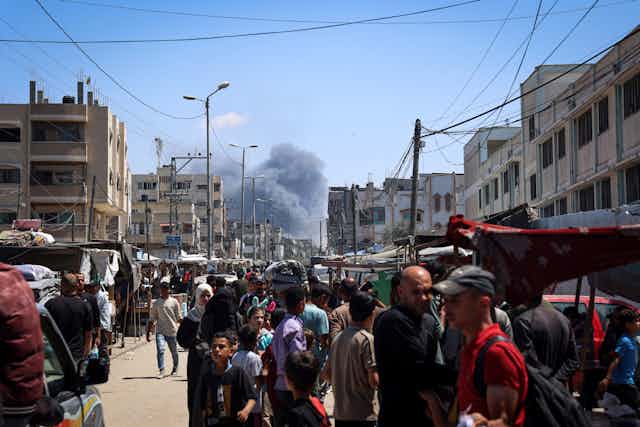 A crowd of people stand in a street that looks packed with traffic. Smoke billows behind them down the street above buildings. 