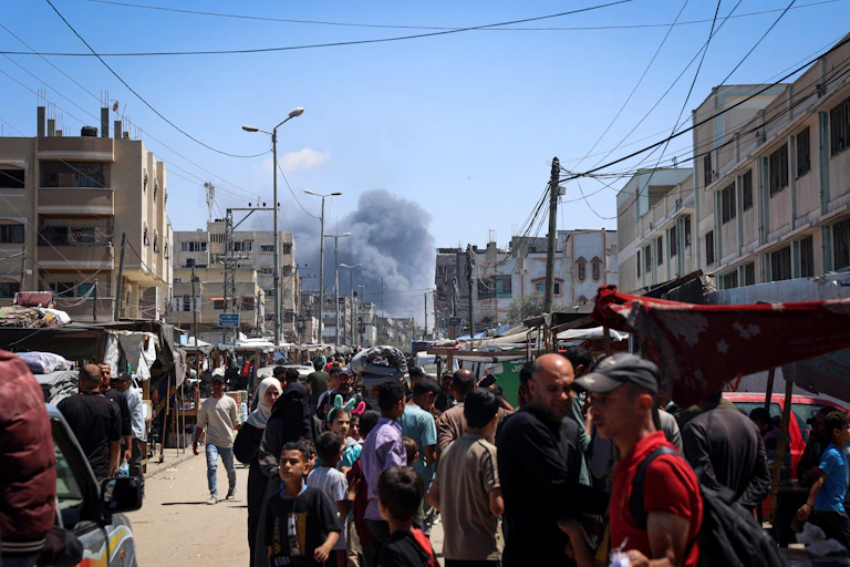 A crowd of people stand in a street that looks packed with traffic. Smoke billows behind them down the street above buildings.