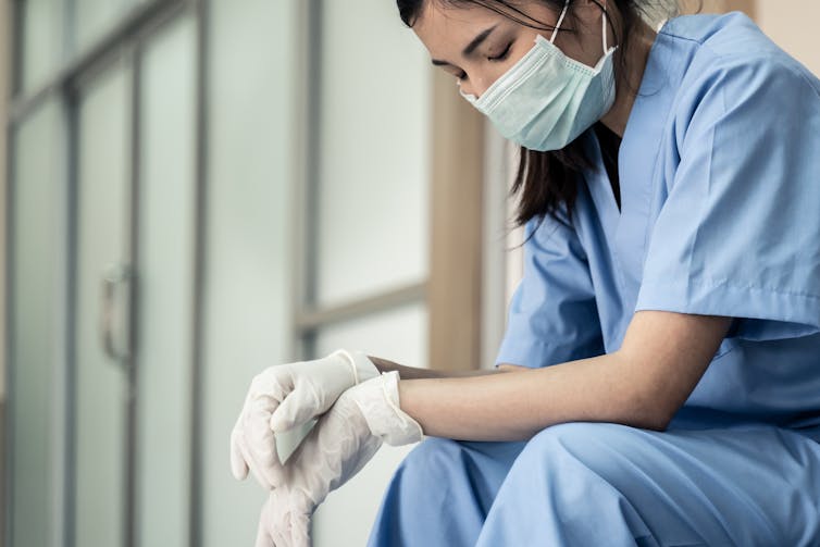 A health-care worker in blue scrubs and a face mask looking tired