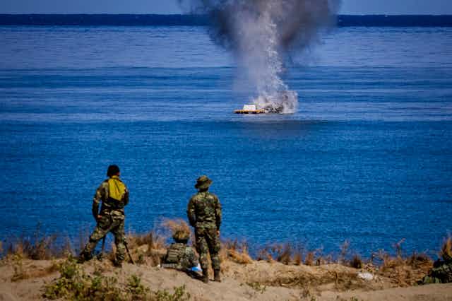Men in fatigues stand and sit on a beach staring at a structure at sea which has smoke billowing from it.