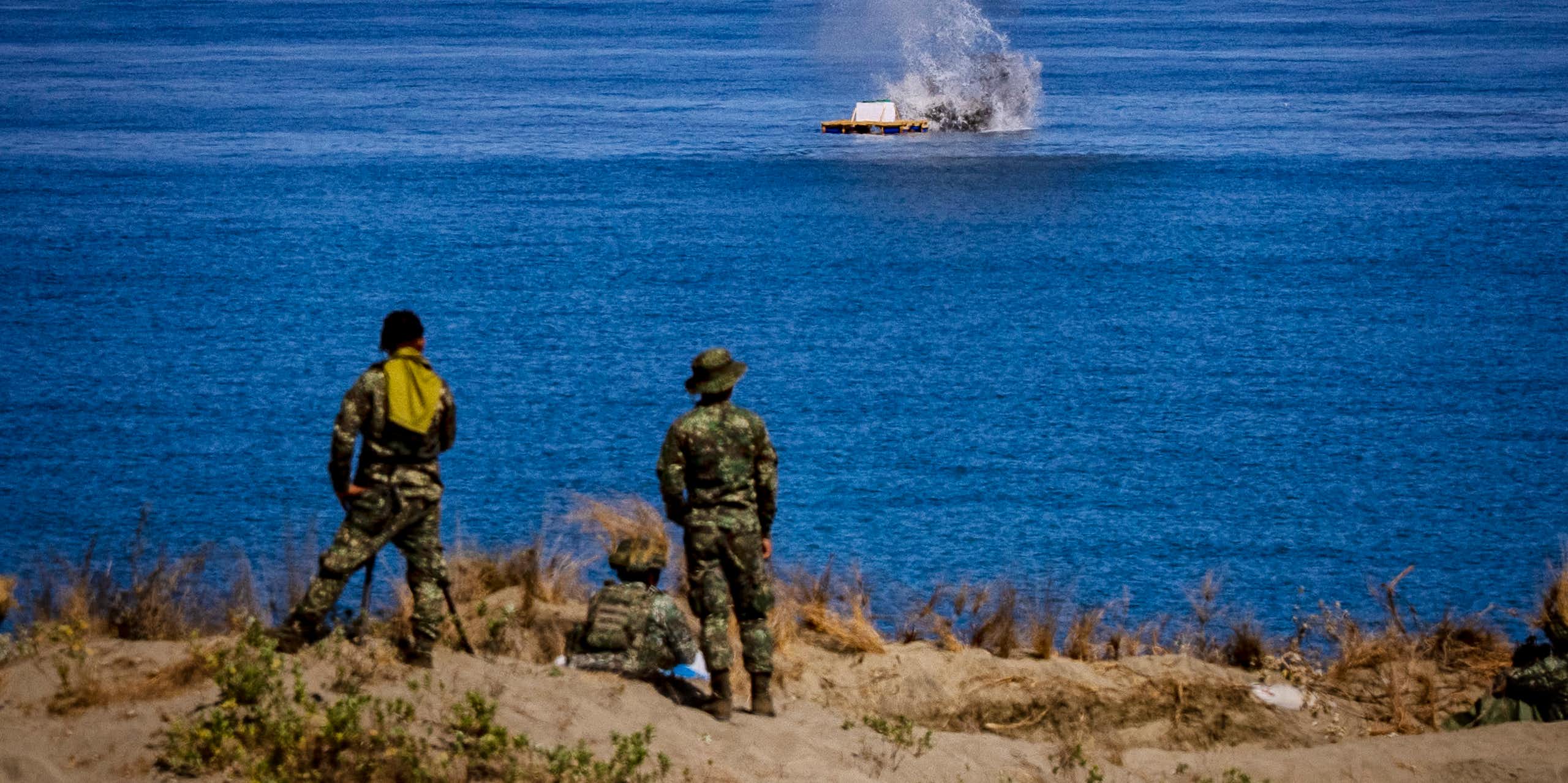Men in fatigues stand and sit on a beach staring at a structure at sea which has smoke billowing from it.