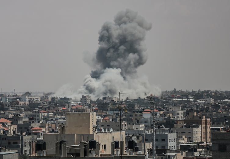 A large plume of smoke rises above many buildings and against a grey sky, seen in the distance.