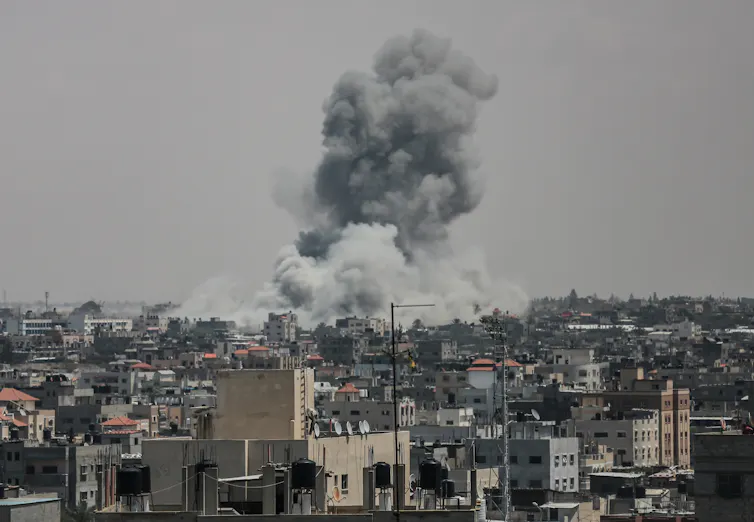 A large plume of smoke rises above many buildings and against a grey sky, seen in the distance.
