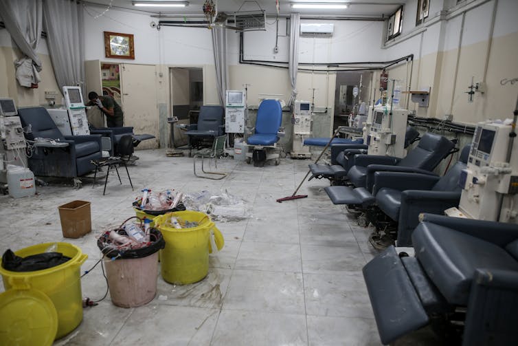 Several blue armchairs and hospital equipment are in a messy looking room, with buckets and plastic medical materials on the ground.