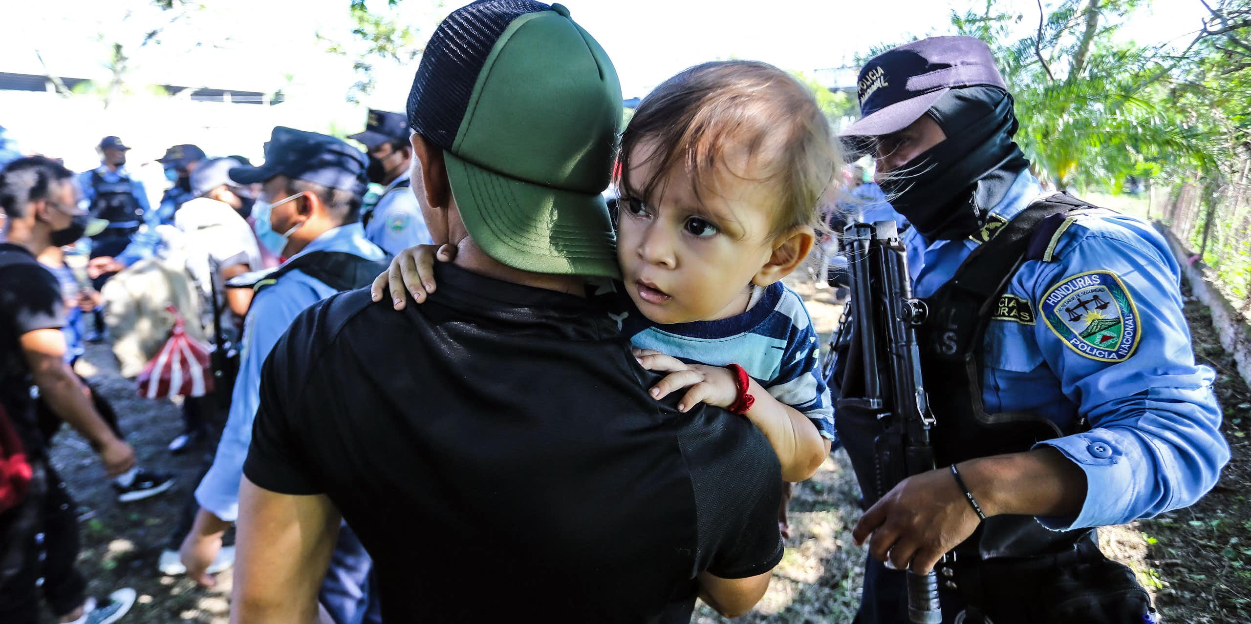 A man in a baseball cap holds a child next to a uniformed man holding a gun.