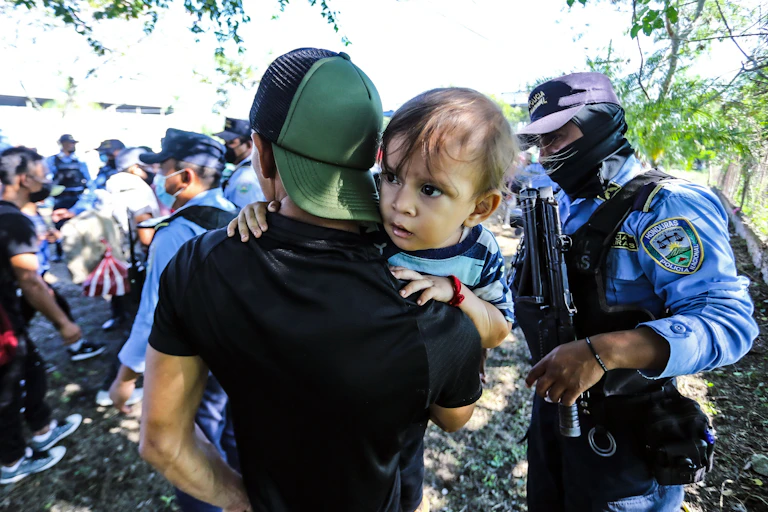A man in a baseball cap holds a child next to a uniformed man holding a gun.