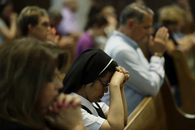 A group of people kneeling in pews in a church, with some holding their hands together in prayer.