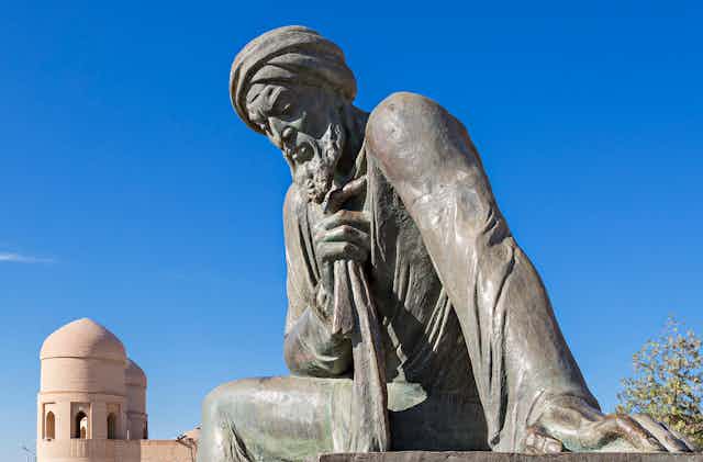 A grey stone figure with chiseled features and a turban set against a blue sky.