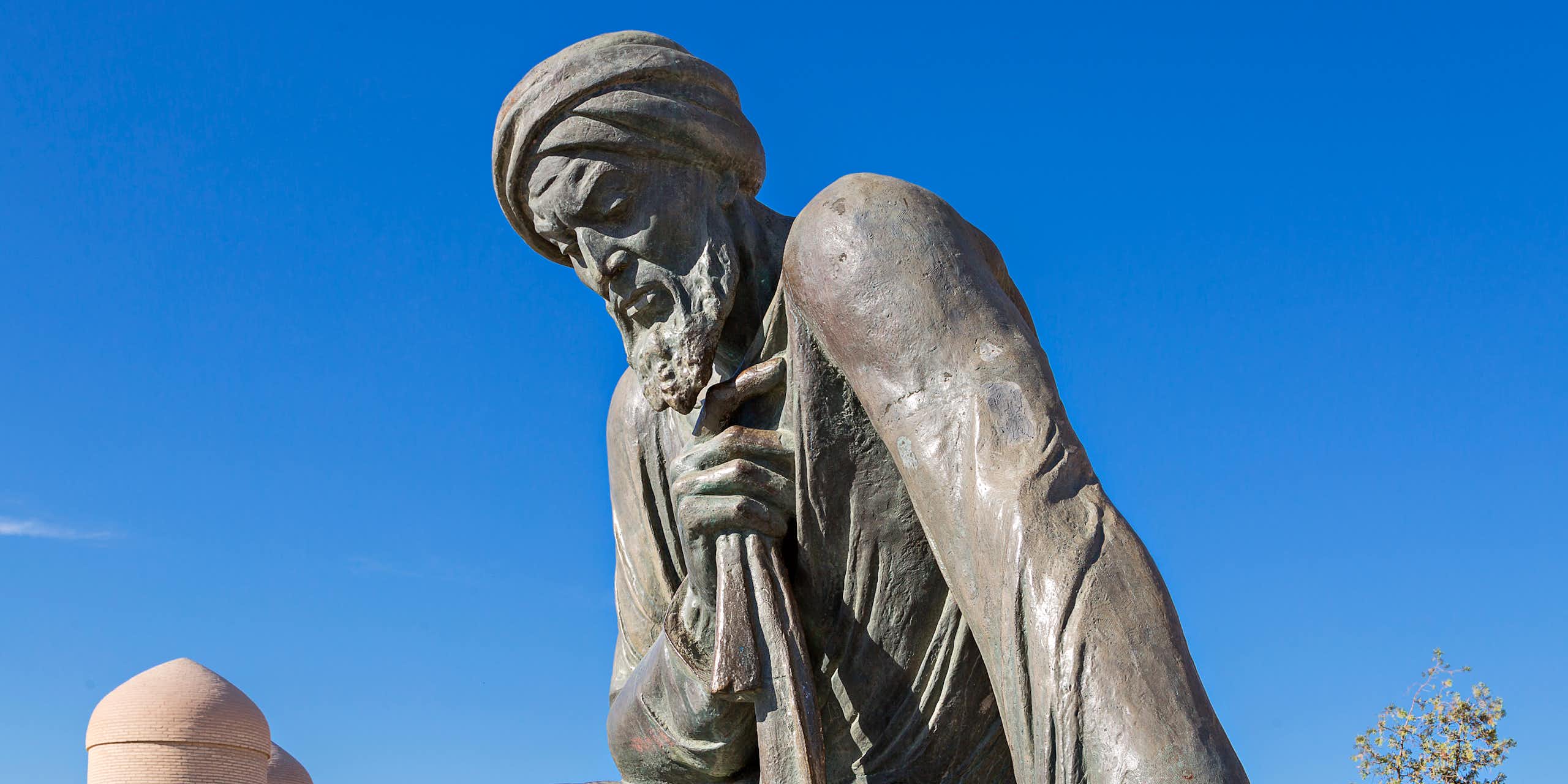 A grey stone figure with chiseled features and a turban set against a blue sky.