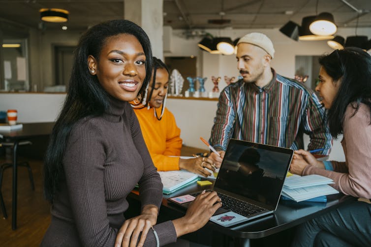 A student smiles while looking at her laptop while others do work at a table.