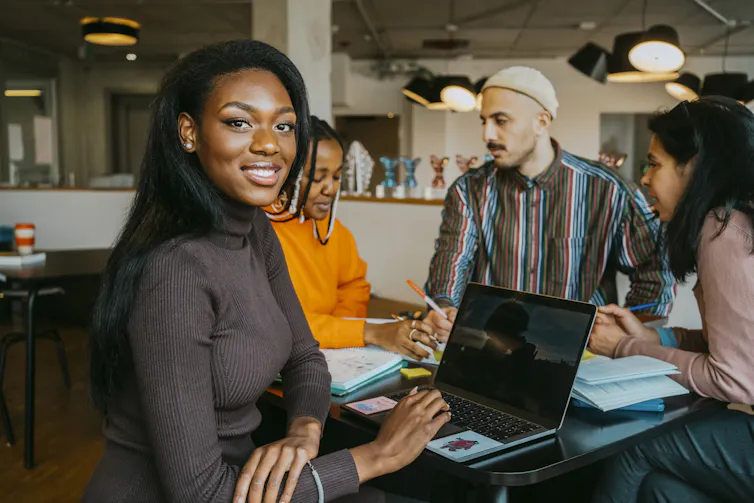 A student smiles while looking at her laptop while others do work at a table.