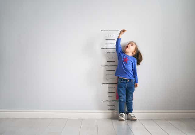 Little girl in cape measuring her height on wall chart. 