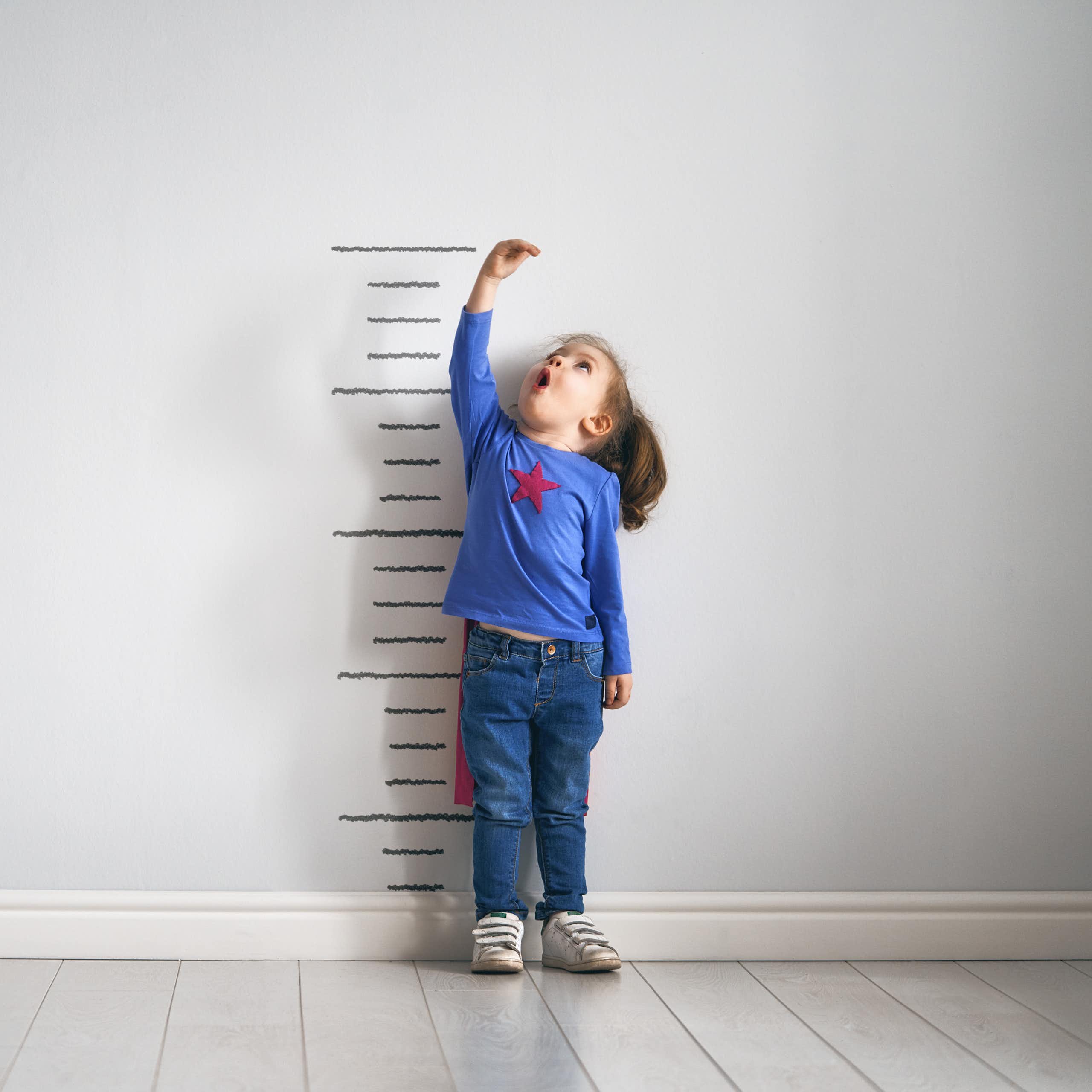 Little girl in cape measuring her height on wall chart. 