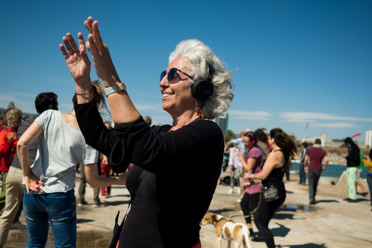 A grey-haired woman dancing on a beach with headphones on smiling and swaying to the music