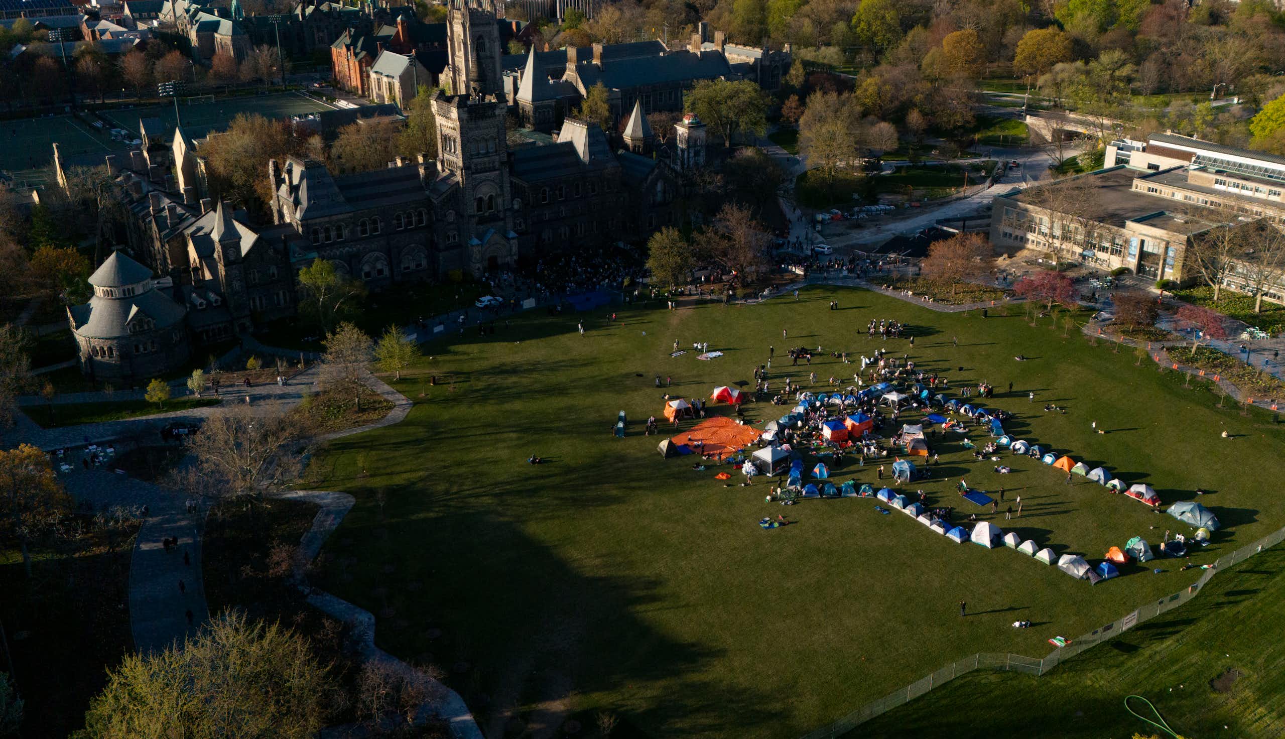 An aerial view shows an encampment on a grassy area with university buildings surrounding it.