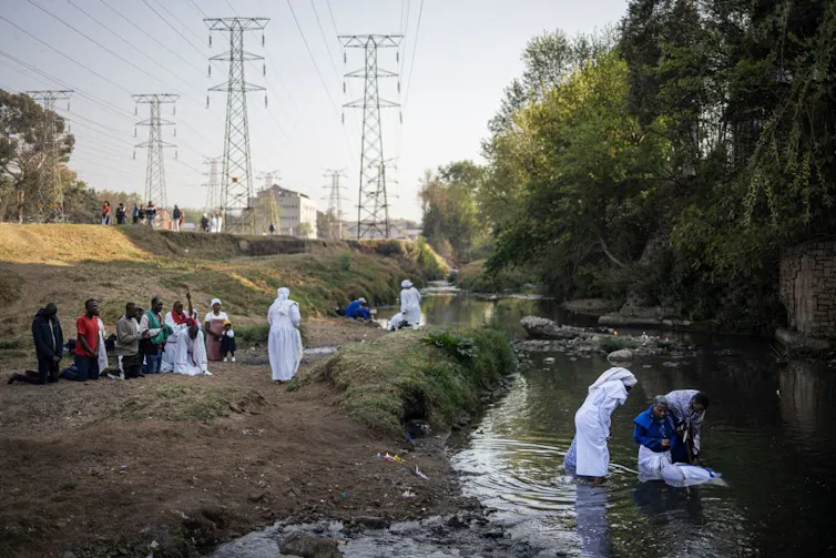 A stream in a field with buildings in the distance. People, some in flowing white robes, gather and watch a baptism in the stream.