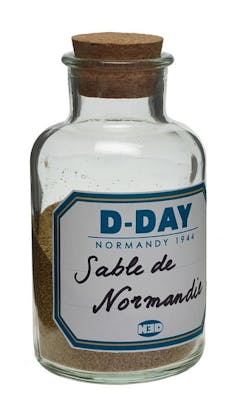 A glass vial holds sand and bears a label reading 'D-Day Sable de Normandie.'