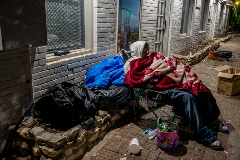 A man wrapped in blankets sits in a chair on a sidewalk near plastic bags and trash.