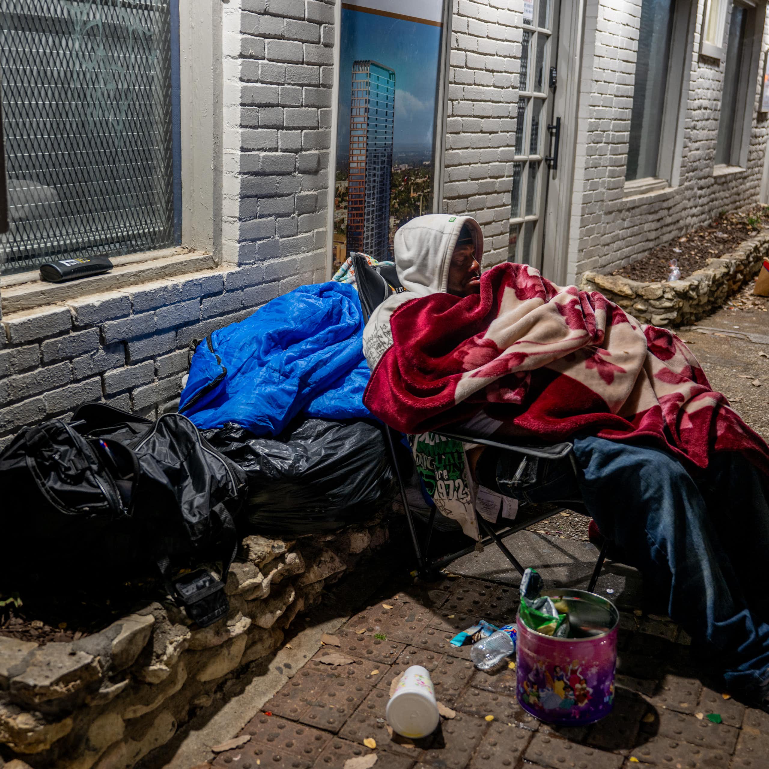 A man wrapped in blankets sits in a chair on a sidewalk near plastic bags and trash.