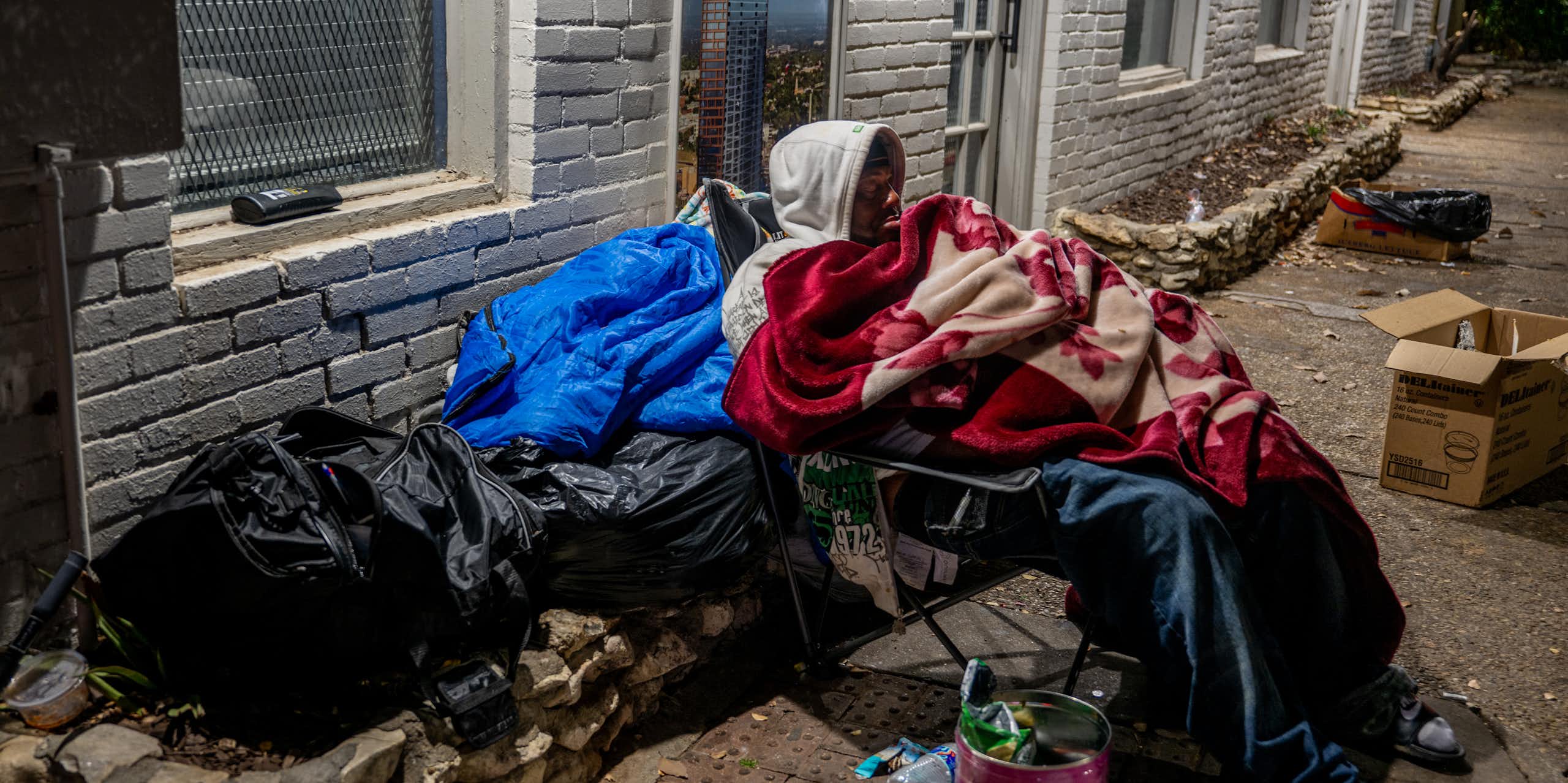 A man wrapped in blankets sits in a chair on a sidewalk near plastic bags and trash.