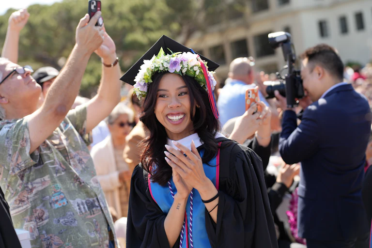 A young woman in a graduation gown and string of flowers around her head smiles as others standing near her take pictures.