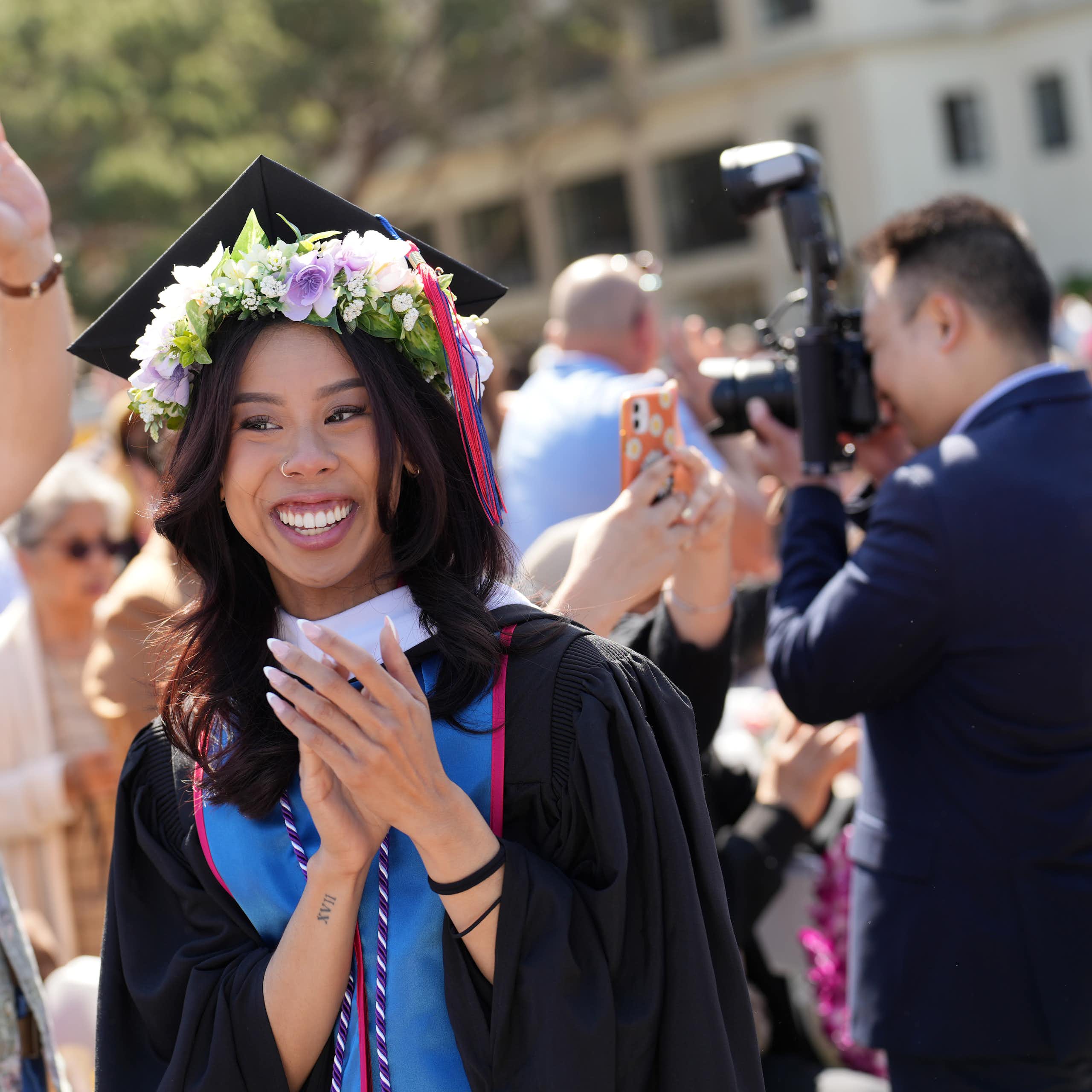 A young woman in a graduation gown and string of flowers around her head smiles as others standing near her take pictures.
