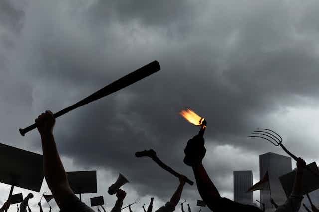 Arms of an angry mob holding protest signs, megaphones, pitchforks, molotov cocktails, and baseball bats, among other weapons